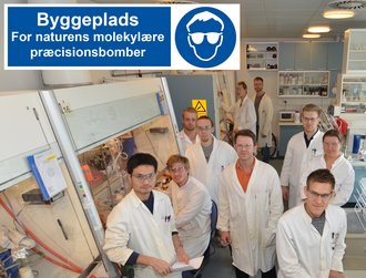 Thomas Bjørnskov Poulsen er manden i midten, iført en orange trøje under kitlen, omgivet af laboratoriets medlemmer. Klik på billedet for at se det i fuld størrelse. Foto: AU